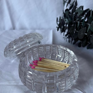 May include: A clear, round glass match holder with a lid, filled with wooden matches with pink tips. The holder has a textured, ribbed design. The lid is off to the side. Decorative greenery is in the background.