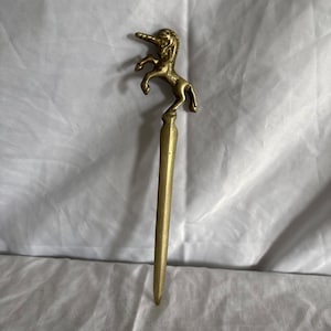 May include: A gold-toned metal letter opener featuring a sculpted unicorn figure at the top. The unicorn is depicted in a rearing position with its front legs raised. The handle transitions into a long, smooth, tapered blade with a pointed tip.