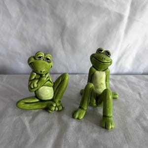 May include: Two green ceramic frog ornaments in a relaxed pose. The frogs have white bellies and expressive eyes. One frog has its arms crossed, while the other sits with its legs extended. A whimsical decorative item.