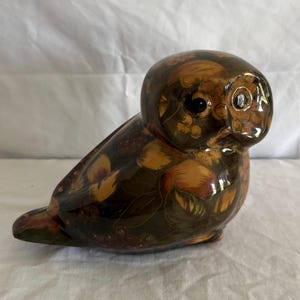 Könnte beinhalten: Dekorative Eulenfigur mit glänzendem Finish. Die Eule ist hauptsächlich dunkelbraun mit einem Muster aus orangefarbenen, gelben und grünen Blumen- und Fruchtdesigns. Die Eule hat große, schwarze Augen und einen runden Kopf.
