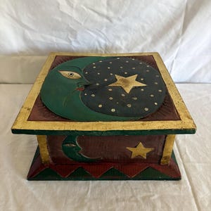 Könnte beinhalten: Eine Holzkiste mit einem himmlischen Thema. Die Kiste zeigt einen grünen Halbmond mit einem Gesicht, einen dunkelblauen Sternenhimmel und einen goldenen Stern. Die Kiste ist mit roten, goldenen und grünen Akzenten bemalt.