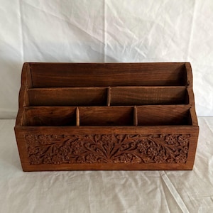 May include: A rectangular wooden desk organiser featuring a dark brown finish and multiple tiered compartments. The front panel is decorated with an intricate, hand-carved floral pattern that spans the entire width of the storage unit.