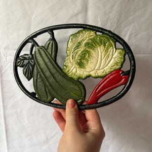 Può includere: Sottopentola in ghisa con design a verdure. La cornice ovale nera contiene zucchine verdi, un cavolo bianco e verde e un peperoncino rosso. Il sottopentola è tenuto da una persona.