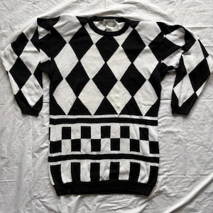 May include: Black and white sweater with a diamond pattern on the upper portion and a checkerboard design on the lower portion. The long-sleeved sweater is made of a soft knit material and is laid flat on a white surface.