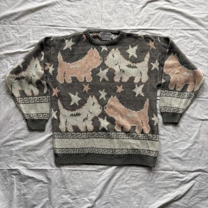 Può includere: Un maglione grigio con un motivo ripetuto di cani dei cartoni animati rosa e bianchi e stelle. Il maglione ha un colletto, polsini e orlo a coste con un motivo a chiave greca. Il marchio "AVOCADO" è visibile sul colletto.