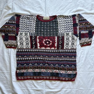 Könnte beinhalten: Ein langärmeliger Pullover mit Patchwork-Design, bestehend aus verschiedenen geometrischen, floralen und abstrakten Mustern in den Farben Burgunderrot, Marineblau, Weiß und Grau. Das Kleidungsstück hat einen Rundhalsausschnitt mit einem Etikett.