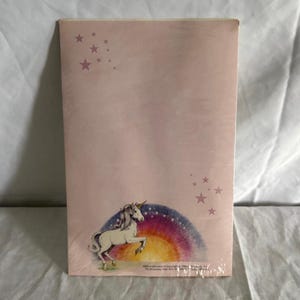May include: A rectangular notepad with a pale pink cover featuring a white unicorn standing in front of a colourful rainbow. Small pink stars are scattered in the top left and bottom right corners. Text at the bottom includes ISBN 0-394-84371-8.