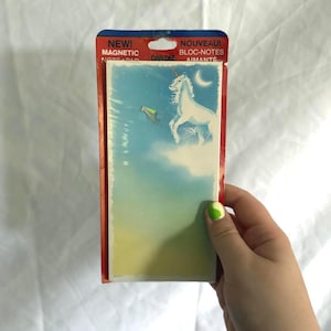 Könnte beinhalten: Ein rechteckiger magnetischer Notizblock in der Originalverpackung. Der Block zeigt einen blauen und gelben Farbverlauf mit einem weißen Einhorn auf einer Wolke unter einer Mondsichel. Die Verpackung trägt die Aufschrift NEW! MAGNETIC NOTE PAD und NOUVEAU! BLOC-NOTES AIMANTÉ.