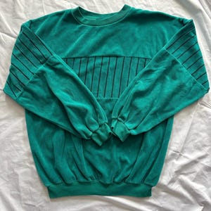 Può includere: Felpa color teal con strisce verticali nere sul petto e sulla parte superiore delle maniche. Le maniche lunghe hanno polsini arricciati. La felpa ha un girocollo e un orlo a coste.