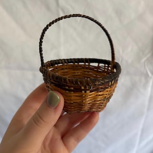 May include: A small, woven basket with a handle. The basket is made of brown and tan wicker, with a darker brown trim around the top. The handle is also made of twisted brown wicker. The basket is held in a hand.