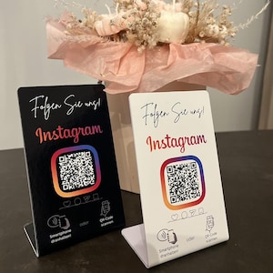 Könnte beinhalten: Zwei kleine, rechteckige Schilder mit dem Text "Folgen Sie uns! Instagram" und einem QR-Code. Ein Schild ist schwarz, das andere weiß. Eine Blumengesteck mit Kürbissen und Trockenblumen befindet sich im Hintergrund.