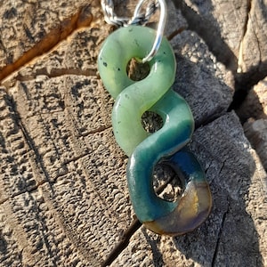 May include: A green and blue glass pendant necklace with a silver chain. The pendant features three interlocking, rounded shapes. The glass has a gradient of colors, transitioning from light green to deep blue. The necklace is displayed on a weathered wooden surface.