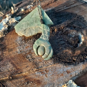 May include: A green jade pendant carved in the shape of a hand holding a sphere. The pendant is on a brown wooden surface.