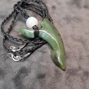 Könnte beinhalten: Halskette mit grünem Jade-Anhänger und schwarzem Band. Der Anhänger ist in Hakenform geschnitzt und hat eine weiße Perle darüber. Die Halskette hat silberne Verschlüsse.