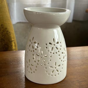 May include: White ceramic oil burner with a floral design. The burner has a wide, round top and a narrow base.
