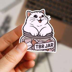 Puede incluir: Una pegatina con un gato de dibujos animados dentro de un tarro con la etiqueta "TBR JAR". El gato es blanco y tiene una expresión feliz. El tarro contiene trozos de papel rosa y marrón.