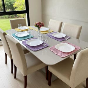 May include: A dining table set for six with beige chairs and a grey tabletop. Each place setting has a white plate on a crocheted placemat in yellow, teal, purple, and pink. Wine glasses and a vase of flowers complete the setting.