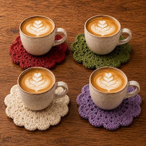 May include: Four speckled ceramic mugs filled with latte coffee featuring heart-shaped foam art, each resting on a round, scalloped-edge crocheted coaster in red, olive green, cream, and purple, arranged in a grid on a dark wood surface.