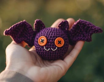 Puede incluir: Un murciélago morado tejido a mano con ojos de botón naranja y una pequeña sonrisa de alambre plateado. El murciélago tiene alas extendidas y orejas pequeñas. Este juguete caprichoso es ideal para Halloween.