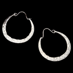 May include: A pair of silver hoop earrings with a hammered texture. The earrings have a simple, classic design with a hook closure. The hoops are round and have a slightly irregular surface, adding visual interest. The background is black.