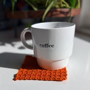 May include: A white ceramic coffee mug with the word "coffee" printed on the side, resting on a small, square, orange knitted coaster. The mug has a handle and a stacked design. The coaster is made of a textured, woven material.