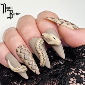 Könnte beinhalten: Ein Set aus langen, mandelförmigen, matten grauen Nägeln mit Nagelkunst zum Thema Schlange. Die Designs umfassen Schlangenköpfe und strukturierte Schlangenhautmuster in Beige- und Brauntönen. Der Name des Künstlers, Naomi Barker, befindet sich oben links.