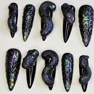 Op de afbeelding: Tien zwarte en iriserende nageltips met verschillende ontwerpen, waaronder slangen, schubben en swirls.