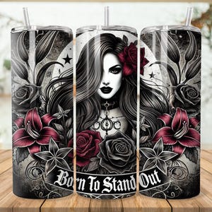 May include: A black and white tumbler with a gothic design featuring a woman with long black hair, red roses, and lilies. The text "Born to Stand Out" is printed on the tumbler.