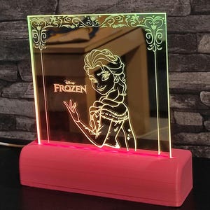 Op de afbeelding: Een roze LED-nachtlampje met een Disney Frozen-ontwerp. Het licht heeft een rechthoekige vorm met een gedetailleerde omtrek van Elsa, het Frozen-personage, en het Disney Frozen-logo. De basis is effen roze.