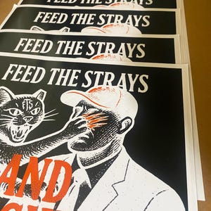 May include: A stack of black and white posters with the text "FEED THE STRAYS" at the top. The bottom of the poster features a graphic of a cat attacking a man. The text "AND FIGHT" is below the graphic. The bottom right corner has the text "TWENTY-FIVE PERCENT OF ALL PROFITS SAVE STREET CATS".
