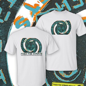 Może przedstawiać: Biały t-shirt z krótkim rękawem z grafiką kociego pyska w okrągłej ramce z napisem "YOUR GOVERNMENT IS LYING" oraz "FEED THE STRAYS" poniżej. Wzór wykorzystuje czarne, turkusowe i pomarańczowe kolory na tkaninie.