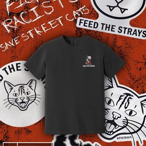 以下が含まれることがあります： 猫のバンダナと「FEED THE STRAYS」の文字が入った小さなグラフィックが特徴の黒いTシャツ。画像には猫をテーマにしたステッカーと「FIGHT RACISTS SAVE STREET CATS」の文字が含まれています。背景はテクスチャ加工されたオレンジ色です。