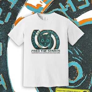 May include: A white short-sleeve t-shirt featuring a graphic of a cat face centered within a teal and orange spiral design. The text "FEED THE STRAYS" is printed below the cat, with additional text surrounding the spiral and at the bottom.