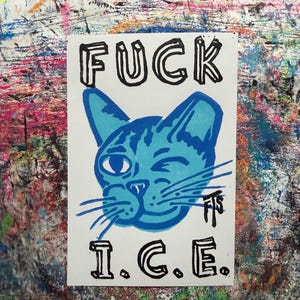 Puede incluir: Una impresión rectangular blanca con una ilustración de un gato azul guiñando un ojo. La impresión incluye el texto "FUCK I.C.E." en negro. El fondo es un patrón abstracto y colorido.