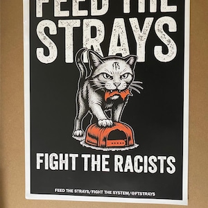 May include: Black and white poster with the text "FEED THE STRAYS" and "FIGHT THE RACISTS". A gray cat with a red object in its mouth stands on an orange bowl. The poster also includes the text "FEED THE STRAYS/FIGHT THE SYSTEM/@FTSTRAYS".