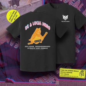 以下が含まれることがあります： 猫のグラフィックがプリントされた黒いTシャツ。1枚のシャツには「BE A LOCAL HERO」と「FEED THE STRAYS」の文字が入った猫のグラフィックが、もう1枚のシャツには猫の顔と「FEED THE STRAYS」の文字が入っています。