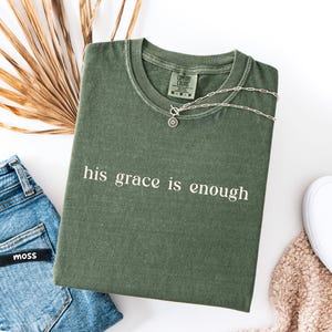 May include: An olive green t-shirt with the phrase "his grace is enough" printed in white. A silver necklace with a circular pendant rests on the shirt. The shirt is paired with blue jeans and a white shoe.