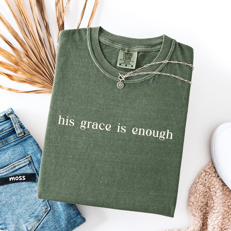 May include: An olive green t-shirt with the phrase "his grace is enough" printed in white. A silver necklace with a circular pendant rests on the shirt. The shirt is paired with blue jeans and a white shoe.