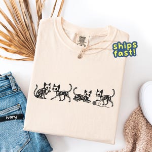 May include: Ivory-colored t-shirt featuring a black skeleton cat design. The design shows four cats in different poses, with one playing with a ball of yarn. The shirt has a crew neck and a tag that says "Comfort Colors".