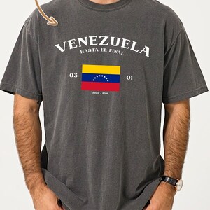 May include: Dark gray t-shirt with the word "VENEZUELA" in white, and the phrase "HASTA EL FINAL" above a Venezuelan flag graphic. The shirt also has the text "03 01" and "2020 - ZAM". The shirt is made of soft cotton.