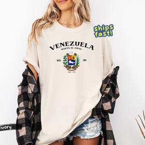 May include: Ivory-colored t-shirt featuring the word "VENEZUELA" above a coat of arms design. The coat of arms includes a horse, wheat, and other symbols. The text "HASTA EL FINAL" is above the coat of arms.