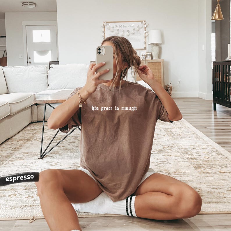 May include: Espresso-colored t-shirt with the text "his grace is enough" in white. The shirt is worn with white shorts and striped socks. The person is sitting on a patterned rug in a living room setting.