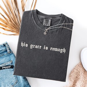 May include: Dark gray t-shirt with the text "his grace is enough" in white gothic font. A silver necklace with a circular pendant rests on the shirt. The shirt is paired with blue jeans and a beige sweater.
