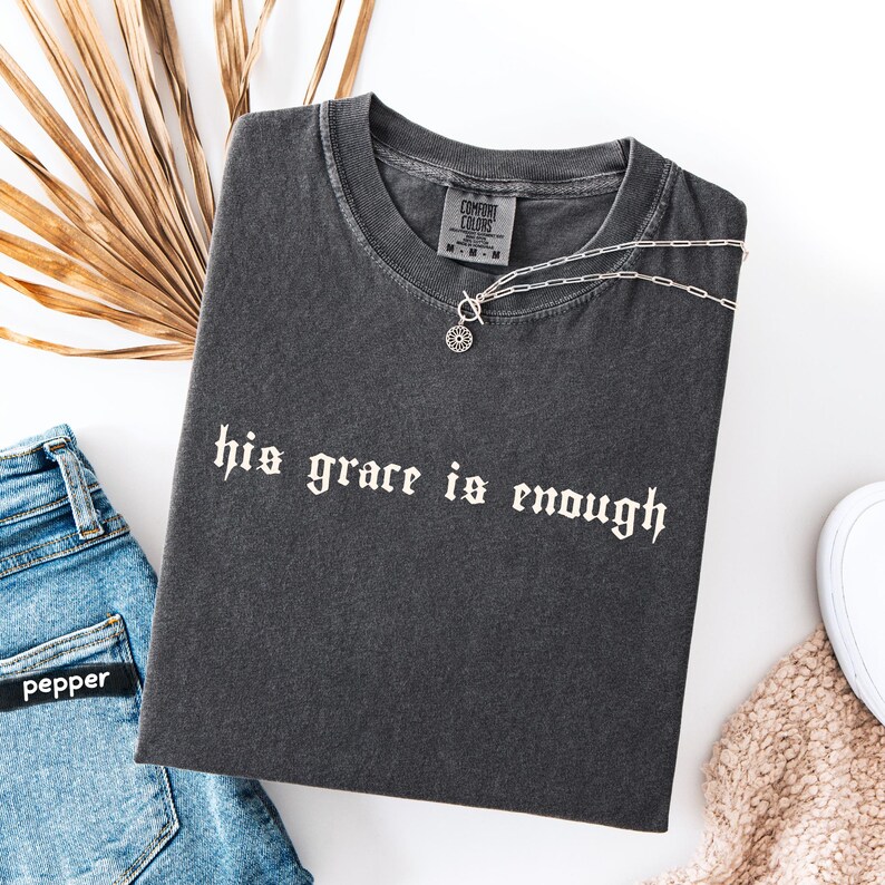 May include: Dark gray t-shirt with the text "his grace is enough" in white gothic font. A silver necklace with a circular pendant rests on the shirt. The shirt is paired with blue jeans and a beige sweater.