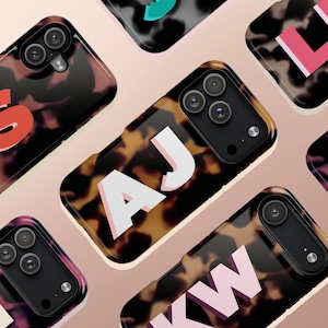 May include: A collection of phone cases with a tortoiseshell pattern. Each case features a different set of white and pink initials. The cases have a black camera module and are arranged on a light pink background.
