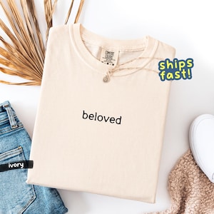 May include: Ivory-colored t-shirt with the word "beloved" printed in black. The shirt is folded and displayed with a pair of blue jeans, a necklace, and a white shoe. The shirt is from Comfort Colors.