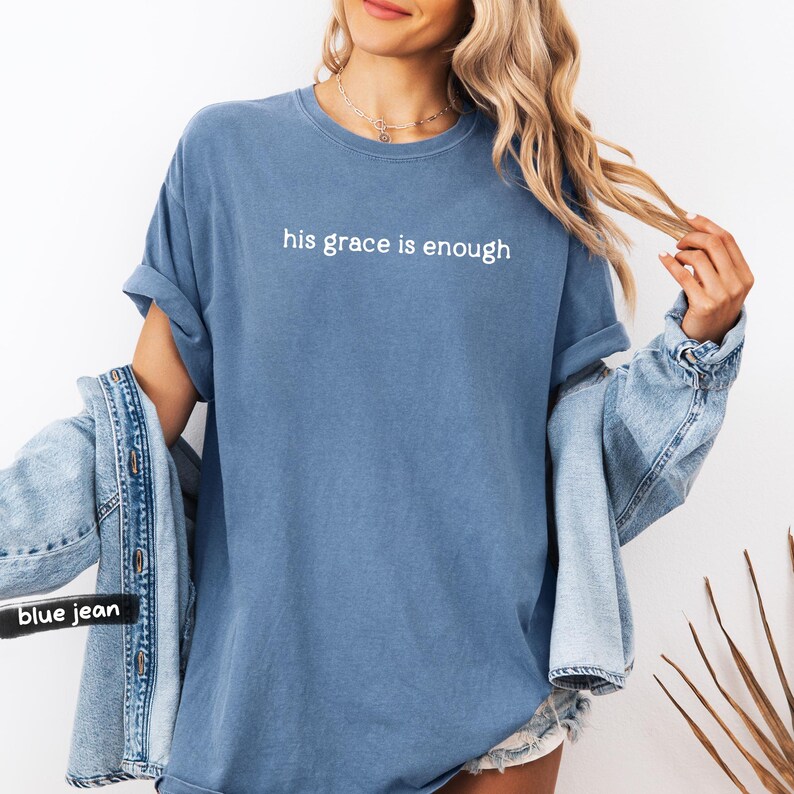 May include: A blue t-shirt with the text "his grace is enough" in white. The shirt has rolled-up sleeves and is paired with a denim jacket. The overall style is casual and comfortable.