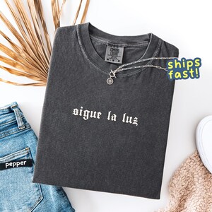 May include: Dark gray t-shirt with the phrase "sigue la luz" in white lettering. A silver necklace with a circular pendant rests on the shirt. The shirt is folded on a white surface next to blue jeans and a beige sweater.