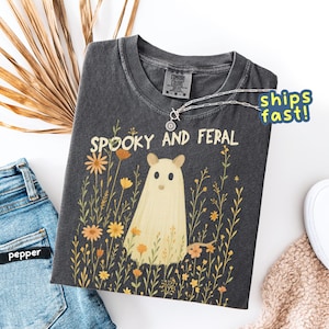 May include: Dark gray t-shirt with the words "Spooky and Feral" and a cartoon ghost design. The ghost is surrounded by yellow and orange flowers. A silver necklace is draped across the shirt.