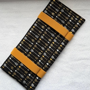 May include: A black and gold fabric wallet with a repeating arrow pattern. The wallet has two mustard-colored elastic bands. The arrows are white and gold. The wallet is rectangular and folded.